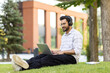 © Tetiana - Young Indian man wearing headphones sitting on the grass outside and smiling while using a laptop he holds on his lap