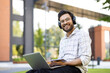 © Tetiana - Portrait of an Indian young man wearing headphones sitting on a bench outside, holding a laptop and a notepad on his lap, smiling and looking at the camera