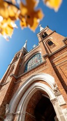 Poster - Magnificent brick church with stained glass window under a clear blue sky framed by golden autumn leaves offering a perspective of history and architectural beauty