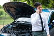 © sorapop - Businessman wearing white shirt and eyeglasses leaning on car, discussing engine problems with roadside assistance on the phone. Driver examining engine with open hood, conveying frustration