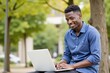 © MT - Smiling young Black man in a blue shirt using a laptop outdoors in a vibrant green park, embodying a relaxed and productive atmosphere in nature.