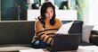 © peopleimages.com - Asian woman, laptop and sofa with documents for finance, budget planning or debt review at home. Female person, computer or paperwork with invoices for financial report, bills or expenses at house