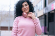 © alfons - Individual portrait of a Caucasian woman wearing a pink hoodie and headphones outdoors. She smiles slightly, standing confidently in an urban environment with a casual and relaxed look.