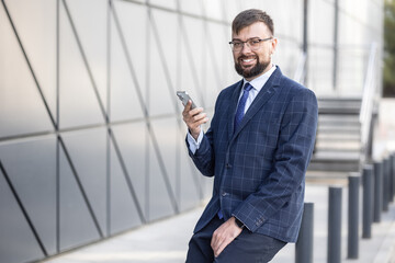 man in collar suit is smiling and holding smartphone in front of building. modern tasks using technology. planning and implementation of work. business trip or checking his messages
