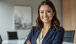© f_bossa - Modern portrait headshot of a friendly ceo executive business worker: A confident businesswoman with dark hair and a warm smile poses with her arms crossed in a modern office setting.