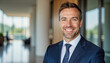© f_bossa - Modern portrait headshot of a friendly ceo executive business worker: A smiling, well-dressed man with short dark hair and a slight beard confidently looks at the camera while standing in a modern,
