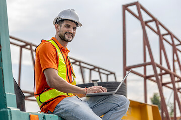 A male civil engineer in orange safety suit is using computer and application running a quality check of precast or cement floor products in a factory with crane background. Technology in a factory