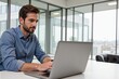 © MT - Young Adult Male Working on Laptop in Modern Office Setting with Large Windows and Cityscape Background