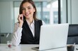 © JustFun - A Young Caucasian Woman in a Black Vest Sitting at a Desk in a Modern Office, Smiling and Engaged in a Phone Call While Working on Her Laptop