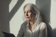 © Isuru - A thoughtful senior woman with gray hair sits by a wall, using a tablet in the sunlight.
