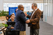 © Drobot Dean - An elderly White man and an adult Black man in business attire talk and hug in an office surrounded by colleagues working at desks. The office includes large windows and potted plants.
