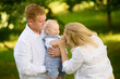 © GRON777 - Dad holds happy baby in arms, during family walk in park, mom tickles child, amuses. Dad and mom play with toddler outside. Family vacation concept, parenthood. International Family Day