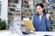 © Liubomir - An excited woman celebrates after reviewing documents in a library setting, surrounded by bookshelves and her laptop.