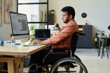 Fototapete - Side view of young multiethnic man with disability sitting in wheelchair and typing on keyboard while using AI image generator