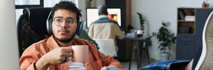 Poster - Header of young restful male office worker in headphones having coffee at break and looking at computer screen while watching tutorial