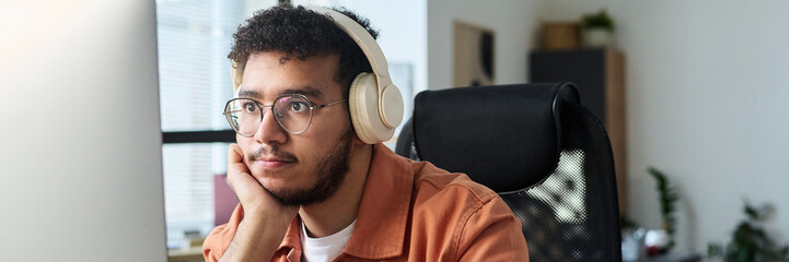 Poster - Header of young serious man in headphones looking at computer screen while watching online video by workplace