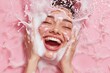 © Init - Young woman with radiant skin joyfully cleanses her face with cleansing foam.  Pink background adds a playful touch.