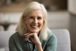 © fizkes - Natural beauty of ages. Portrait attractive positive older woman on retirement with blond hair wrinkled skin sitting on couch with shiny toothy smile looking forward touching chin satisfied with life