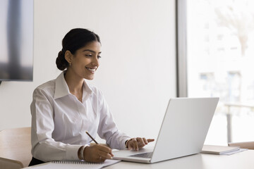 attractive smiling indian woman sitting at table with laptop, holds pen, jotting information, making
