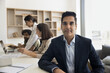 © fizkes - Middle-aged Indian businessman in formal suit looks at camera in office with staff members in background engaged in discussion. Profile picture of project leader, male company boss, executive manager