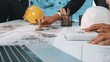 © InfiniteFlow - A focused team of engineers collaborates on detailed construction plans in an office environment, using safety helmets and modern technology for effective project execution. SACTR
