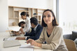 © fizkes - Middle-aged woman looking at camera seated at conference table in office with colleagues in background engaged in discussion. Portrait of project leader, company boss, business executive, authority