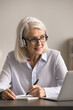 © fizkes - Third age university student. Smiling interested older woman enjoy e-learning with headphones on listen to video lecture on notebook meet teacher online for consultation by videocall take brief notes