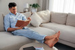 © New Africa - Man reading book on sofa in living room