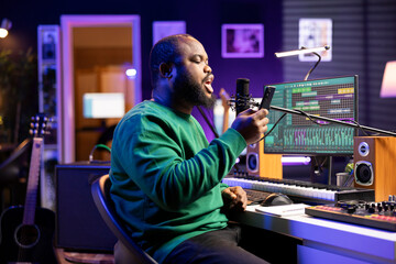 Poster - Skilled musician recording a new song written by him in home studio, reading lyrics from his phone and singing on microphone. Music engineer prepares to edit the track in post production.