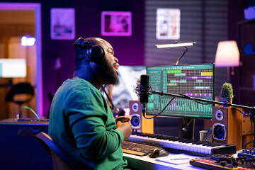 Fototapete - Male musician recording a new song on microphone in home studio, working on editing the recorded tunes in post production. Skilled composer artist performing acoustics on stereo gear.