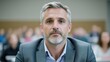 © KUTIL KURO - A high-resolution photo shows a middle-aged man with graying hair, wearing a gray suit. Hes centrally positioned against a blurred background of people. The image is well-lit, with a neutral tone. T