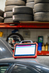 Digital diagnostic scanner tablet on vehicle in auto repair garage with extensive tire storage rack visible. Professional automotive diagnostic tool used in modern car service workshop with organized 