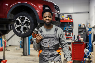 Fototapete - Portrait of young dedicated black auto mechanic standing at workshop with drill in hands and looking at camera.