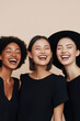 © Sergey - Three diverse women laughing together in stylish black outfits against a neutral background.