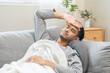 © KMPZZZ - Sick, influenza asian young man, male have fever hand touching forehead for check measure body temperature, illness while lying rest on sofa at home. Health care on virus person.