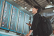 © oatawa - Happy asian man traveller using smartphone checking flight schedule departures board in departure airport terminal hall in front of check in counters. Tourist journey trip concept