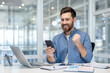 © Liubomir - A happy businessman celebrates success, looking at his phone in the office. There is a laptop, tablet, and documents on the desk.