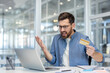 © Liubomir - A frustrated businessman in a modern office, holding a credit card and staring at his laptop with an unhappy expression.