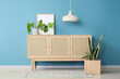 © Pixel-Shot - Chest of drawers with houseplants, lamp and frame near blue wall in room