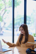 © Jirapong - Smiling young woman engaged in a joyful online e-learning video call, gestu while holding a book with headphones on a wooden table nearby