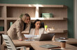 © My Ocean studio - Two professional women are engaged in a business discussion in an office. One is seated at a desk, working on a laptop, while the other, standing, points to documents in her hands.