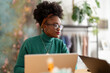 © Studio Marmellata - A young woman with curly hair, glasses, and a green sweater is focused on her laptop while working in a modern office space with a clothing rack and plants in the background.