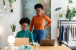 © Studio Marmellata - Two women with curly hair, glasses, and stylish outfits share a friendly conversation while working on laptops at a shared office desk in a bright and modern workspace.
