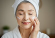 © Laxmico - タオルを巻いて朝のスキンケアを楽しむシニア女性