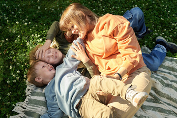 Wall Mural - High angle view of smiling young mother cuddling with two young boys while lying on picnic blanket on green grass in park