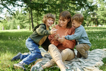 Wall Mural - Candid scene of two young boys giving flowers to mom while playing together on picnic blanket in green park