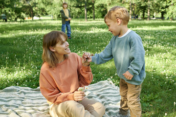 Wall Mural - Candid scene of cute little boy giving flower to mother both smiling happily in green park