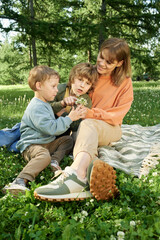 Wall Mural - Vertical full length portrait of happy mother with two young boys sitting on picnic blanket in green park and holding wildflowers