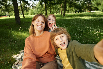 Wall Mural - POV portrait of happy mother and two boys taking selfie photo together while sitting on picnic blanket in green park