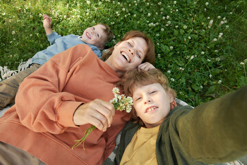 Wall Mural - High angle POV portrait of happy mother and two boys taking selfie photo together while lying on picnic blanket in green grass
